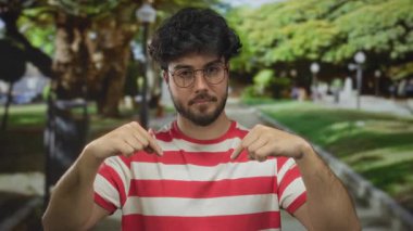 Sakallı ve gözlüklü genç İspanyol bir adam güneşli bir parkta dikilirken yeşil çizgili bir gömlek giyiyordu. Etrafı yemyeşil, açık havayı çağrıştırıyordu..
