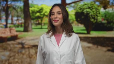 Laboratuvar önlüğü giyen bir kadın güneşli bir parkta arka planda yemyeşil bir yeşillik içeren olumlu ve profesyonel bir hava yayarak başparmağını kaldırıyor..