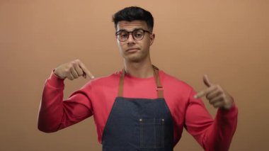 Gözlüklü, kırmızı tişörtlü ve kot önlüklü genç adam kahverengi arka planda kendine işaret ediyor..