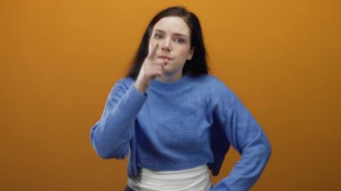 Dışarıdaki kadın canlı turuncu arka plana karşı hareketli ifade veriyor, mavi kazak giyiyor, duygularını vurguluyor, kendinden emin bir şekilde dışarıya işaret ediyor, hoşnutsuzluğunu dile getiriyor..