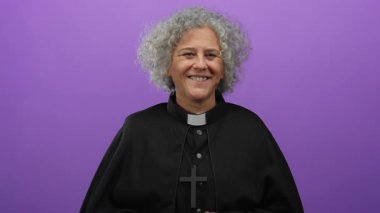 Siyah papaz kıyafeti giymiş gri saçlı yaşlı kadın sırıtışına işaret ederken gülümsüyor, canlı mor bir duvara yaslanıyor, neşeli ve cana yakın bir tavır sergiliyor..