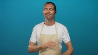 Uzun saçlı sarışın adam kollarını kavuşturuyor ve mavi stüdyoda gözleri kapalı gülümsüyor..