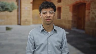 Mavi gömlekli genç İspanyol adam elleri tuğla bina cephesinin dışında dar kaldırım taşı sokakta elleri ve ağzını çerçeveler; sürpriz.