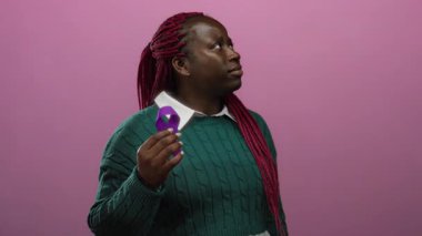 Mor kurdeleli kadın, pembe arka plana karşı düşünceli görünüyor, yeşil bir kazak giyiyor, farkındalığı ve desteği temsil ediyor..