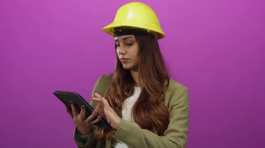 Genç kadın mimar, sarı bir başlık takarak, modern mühendislik ve profesyonel odaklanmayı yansıtan, yalıtılmış, pembe bir arkaplan üzerine kurulmuş bir tablet kullanıyor..