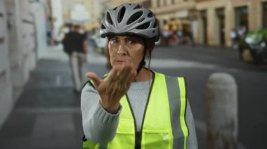 Yoğun bir şehir caddesinde dikilirken yansıtıcı yelek ve kask takan kıdemli kadın, açık hava kentsel güvenlik mesajı iletiyor..