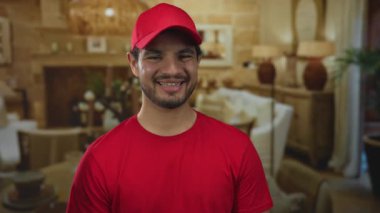 Kırmızı şapkalı ve kırmızı tişörtlü İspanyol genç adam, oturma odası ortamında sıcak bir şekilde gülümsüyor. Mutluluk..