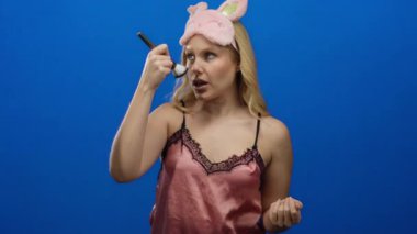 Pembe gece elbisesi ve uyku maskesi takan bir kadın canlı mavi bir arka plan önünde makyaj yapıyor. Güzellik ve rahatlama temalarını gözler önüne seriyor..