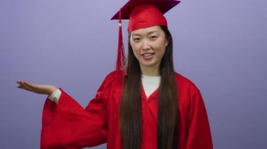 Kırmızı şapkalı ve cüppeli mezun bir kadın. Mor bir arka planda el kaldırılmış, izole stüdyo ortamındaki başarısını ve başarısını kutluyor..