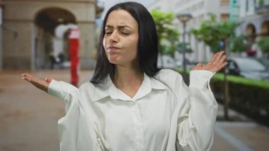 Beyaz tişörtlü kadın, kalabalık bir şehir caddesinde elleri havada, şehir ortamında kargaşa ve belirsizlik içinde dikiliyor..