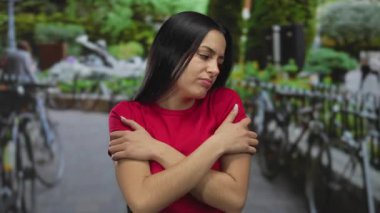 Kırmızı tişörtlü İspanyol kadın, bulanık yeşillik ve bisikletlerle soğuk bir park ortamında kendini kucaklıyor. Soğuk bir rahatsızlık ve yalnız bir iç gözlem taşıyor..