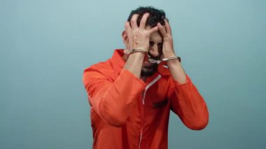 Tutuklu adam turuncu tulum ve kelepçe takıyor, mavi arka planda belirgin bir şekilde jest yapıyor hapsetme ve duygu tasvir eden dramatik bir sahnede.