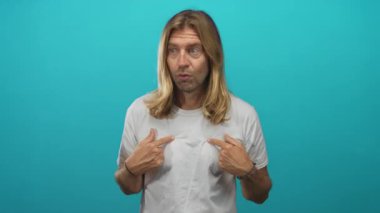 Sarışın, uzun saçlı, turkuaz bir stüdyo stüdyosunda parmaklarını göğsüne gösteren, şaşırmış bir yüz ifadesi olan, sürpriz kişilik keşfi yapan bir adam..