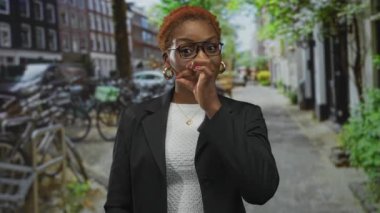 Gözlüklü ve siyah ceketli kadın, kalabalık şehir caddesinde bisiklet parkının ortasında ağzını çimdikliyor; sessizlik.