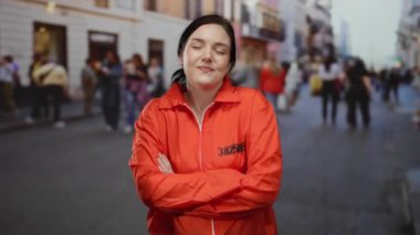 Turuncu hapishane tulumlu esmer kadın kalabalık bir caddede kollarını kavuşturmuş, güvenli bir şekilde dışarıda duruyor..