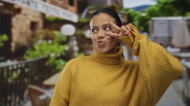 Terasta hardal süveter giymiş bir kadın arka planda bulanık bir açık hava restoranıyla barış işareti sergiliyor. Büyüleyici bir İspanyol atmosferi olduğunu gösteriyor..