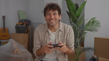 Yeni evde oyun oynayan gülümseyen bir adam. Etrafı karton kutular ve bitkilerle çevrili. Dolaşmayı ve rahatlığı resmediyor.
