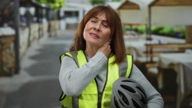 Kasklı ve yansıtıcı yelekli İspanyol bir kadın şehir terasında boynuna dokunuyor. Ağrı ve yorgunluk hissi uyandırıyor..