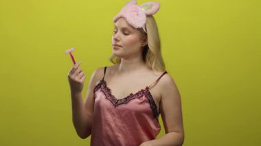 Pembe gecelikli kadın parlak sarı bir arka plana jilet tutuyor, kadınsı ve özenli bir karışım yayıyor, gece temasını tamamlayan neşeli bir göz maskesi takıyor..