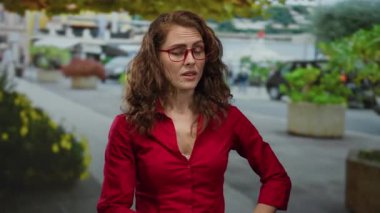 Gözlüklü ve kırmızı tişörtlü bir kadın, yeşil ve park etmiş arabalarla çevrili kalabalık bir şehir caddesinde güpegündüz duruyor..