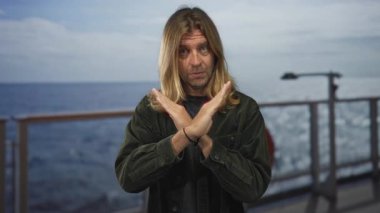 Sarışın ve uzun saçlı bir adam açık denize tepeden bakan güvertede x hareketiyle kollarını kavuşturuyor..