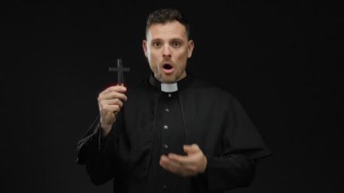 Rahip gibi giyinmiş bir adam karanlık bir arka plana karşı elinde haç, ağzının üzerinde bir el, sürpriz ya da şok, izole edilmiş ve gizemli dini imgeler iletiyor..