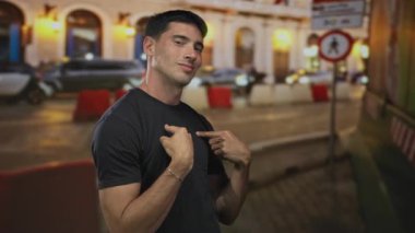 Siyah tişörtlü adam neon ışıklarının altında alacakaranlıkta hareketli tarihi caddede parmak göğse işaret ediyor..