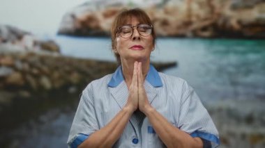 Gözlüklü, çizgili üniforma giyen yaşlı bir kadın deniz kenarındaki bir yerde elleri ve gözleri kapalı, derin derin düşüncelere dalmış ve huzurlu bir şekilde duruyor..