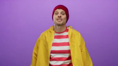 Sarı yağmurluk giyen, çizgili gömlekli genç adam canlı mor bir arka plana karşı jestler yapıyor..