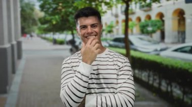 Çizgili tişörtlü genç bir adam şehir caddesinde gülümsüyor, yeşilliklerle çevrili, dışarıda çekici bir şehir portresi sunuyor..