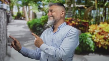 Sakallı orta yaşlı bir adam yeşil yapraklar ve taş duvarlar arasında bir orman yolunda iki parmağını işaret ediyor..