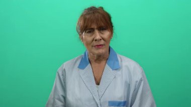 Kıdemli İspanyol kadın temizlikçi yeşil arka plana karşı duruyor çizgili üniforma giyiyor profesyonelliği ve iş ortamını gösteriyor..