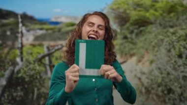 Açık hava parkında yeşil süveter giyen genç bir kadın elinde öğrenci tabelası ve arka planda doğa ile heyecan gösteren bir kadın..