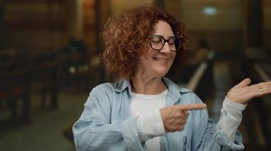 Gülümseyen ve el kol hareketi yapan bir kadın kilisenin arka bahçesinde günlük kıyafetlerle, kapalı bir dini ortamda güven ve pozitiflik gösteren bir tabela..