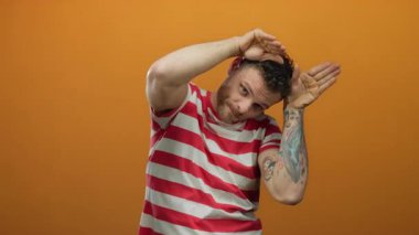 Çizgili tişörtlü genç İspanyol, izole edilmiş turuncu arka plana karşı neşeli kulak hareketi yapıyor..
