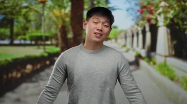 Gri kazaklı genç adam canlı bir park ortamında dikiliyor, yemyeşil ve renkli çiçeklerle çevrili..