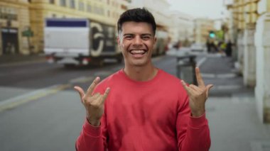 Kırmızı süveter giyen genç adam güler yüzlü ve hareketli bir şehir caddesinde gün ışığında coşkuyla el kol geziyor..