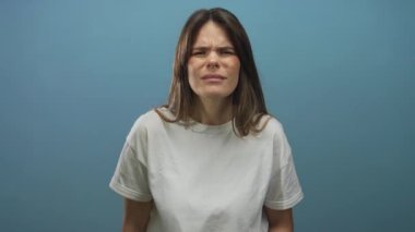 Düz mavi bir arkaplanda beyaz tişört giyen bir kadın, kapalı mavi bir portre ortamında şaşkınlık ya da şaşkınlık belirtileri gösteriyor..