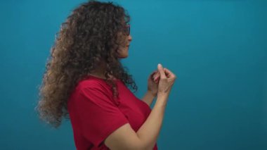 Kırmızı gözlüklü ve gömlekli bir kadın. Stüdyoda mavi bir duvara karşı elleri ve kolları açık bir şekilde sunuluyor..