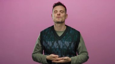 Genç İspanyol bir adam, yeşil bir süveter giyerken sakin ve odaklanmış bir şekilde, izole edilmiş pembe bir arka plan önünde meditasyon yapıyor.
