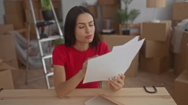 Kırmızı tişörtlü kadın, kutularla çevrili yeni taşınmış bir dairede, etrafındaki eşyalarla birlikte rahatlamış ama odaklanmış bir ifadeyi gösteren belgeleri incelikle tutuyor..