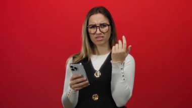 Kırmızı arka planda kafası karışık bir ifadeyle akıllı telefon tutan sarışın kadın, gözlük takıyor ve günlük kıyafetler giyiyor..
