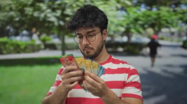 Sakallı genç adam, yeşil doğayla çevrili bir parkta gündüz vakti açık havada gözlük ve çizgili gömlek giyerek İsviçre frangı tutuyor.