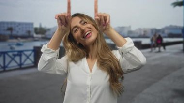 Genç sarışın kadın marina gezinti güvertesinde gün ışığında demirlemiş teknelere el hareketi yapıyor, sevgi dolu neşe.