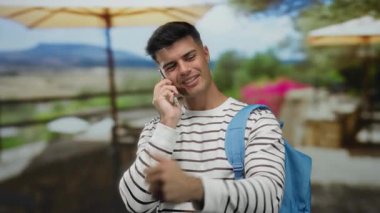 Telefon görüşmesi yapan genç adam şehir terasında duruyor, çizgili gömlek ve sırt çantası giyiyor, restoranın arka planı bulanık ve rahat atmosferi gösteriyor..