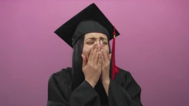 Latin kökenli genç bir kadın neşeyle gülüyor, siyah bir kep ve cübbe giyiyor, parlak pembe bir arka plana işaret ediyor..