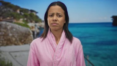 Pembe bornozlu bir kadın deniz kenarında turkuaz su ve kayalarla hayal kırıklığına uğramış bir halde, genç bir İspanyol kadınla açık deniz sahnesini çekiyor..