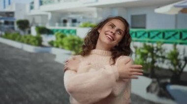 Kadın, arka planda bulanık binalar ve yeşilliklerle aydınlık bir caddede kendine sarılırken sıcak bir şekilde gülümsüyor..