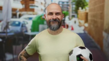 Futbol topu tutan kel bir adam şehir terasında kendine güveniyor, dövmelerini gösteriyor ve sıradan bir şehir manzarasında gülümsüyor..