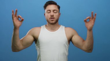 Sakallı genç adam, beyaz atletle meditasyon yapıyor mavi arka planda, gözleri kapalı, durgun pozda.